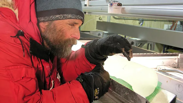 A polar scientist in a red parka examining a cylindrical ice core on a backlit table