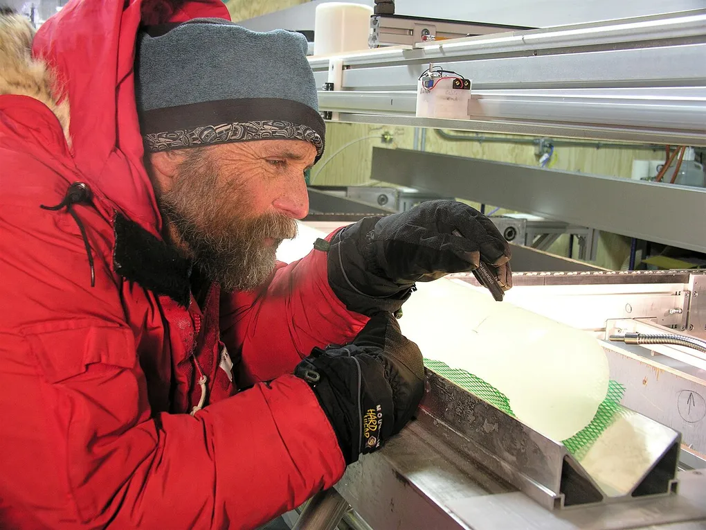 A polar scientist in a red parka examining a cylindrical ice core on a backlit table
